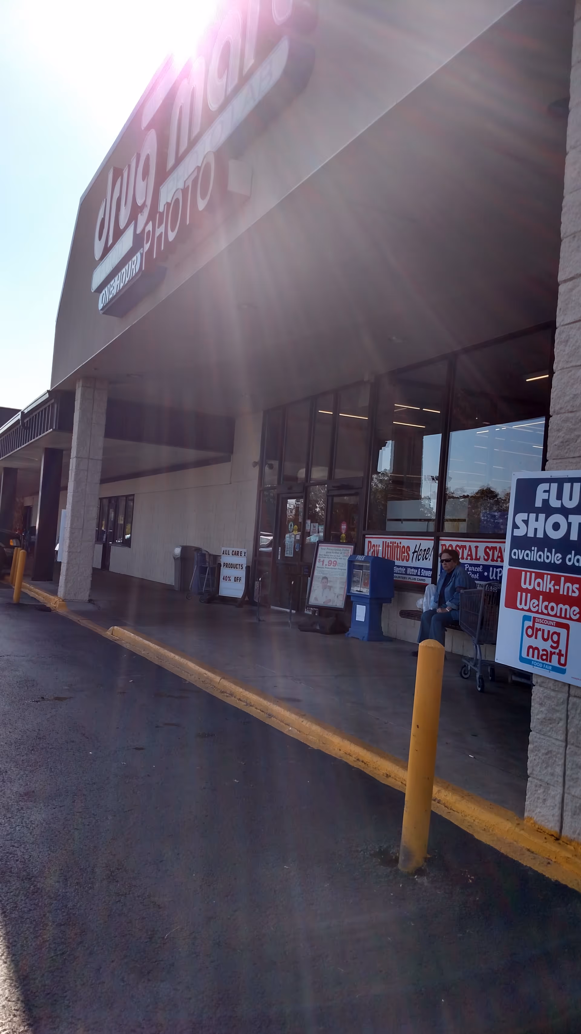 Exterior view of a Discount Drug Mart storefront with sidewalk and a person seated outside.