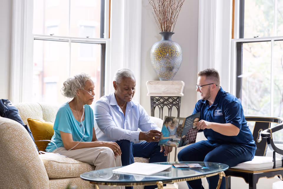 Three adults and a caregiver sit on a living room couch looking at photos together.