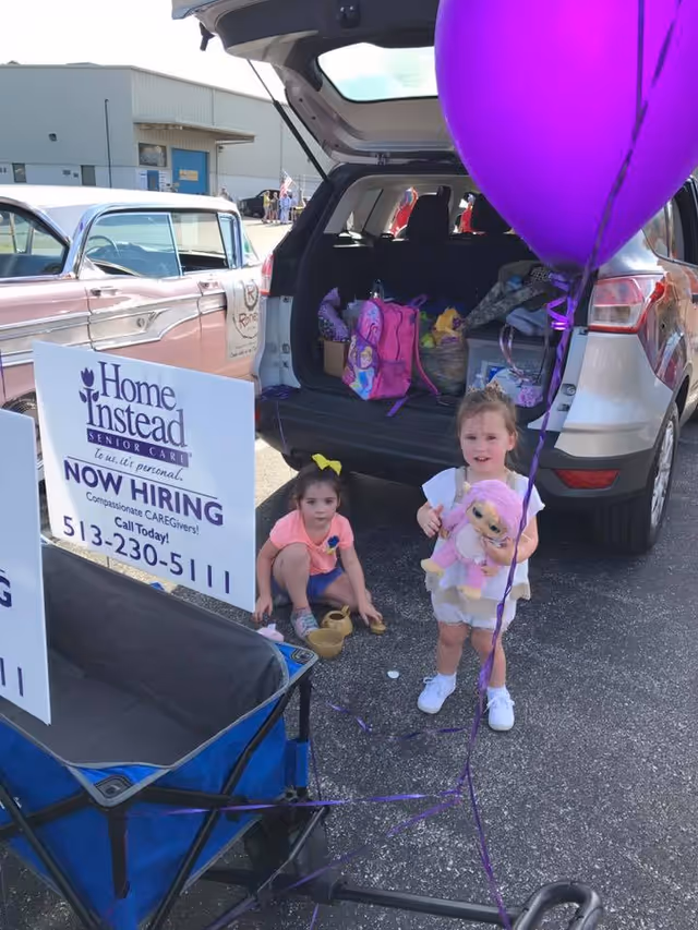 Open SUV trunk at an outdoor Home Instead hiring event, with signs, balloons, and two children.