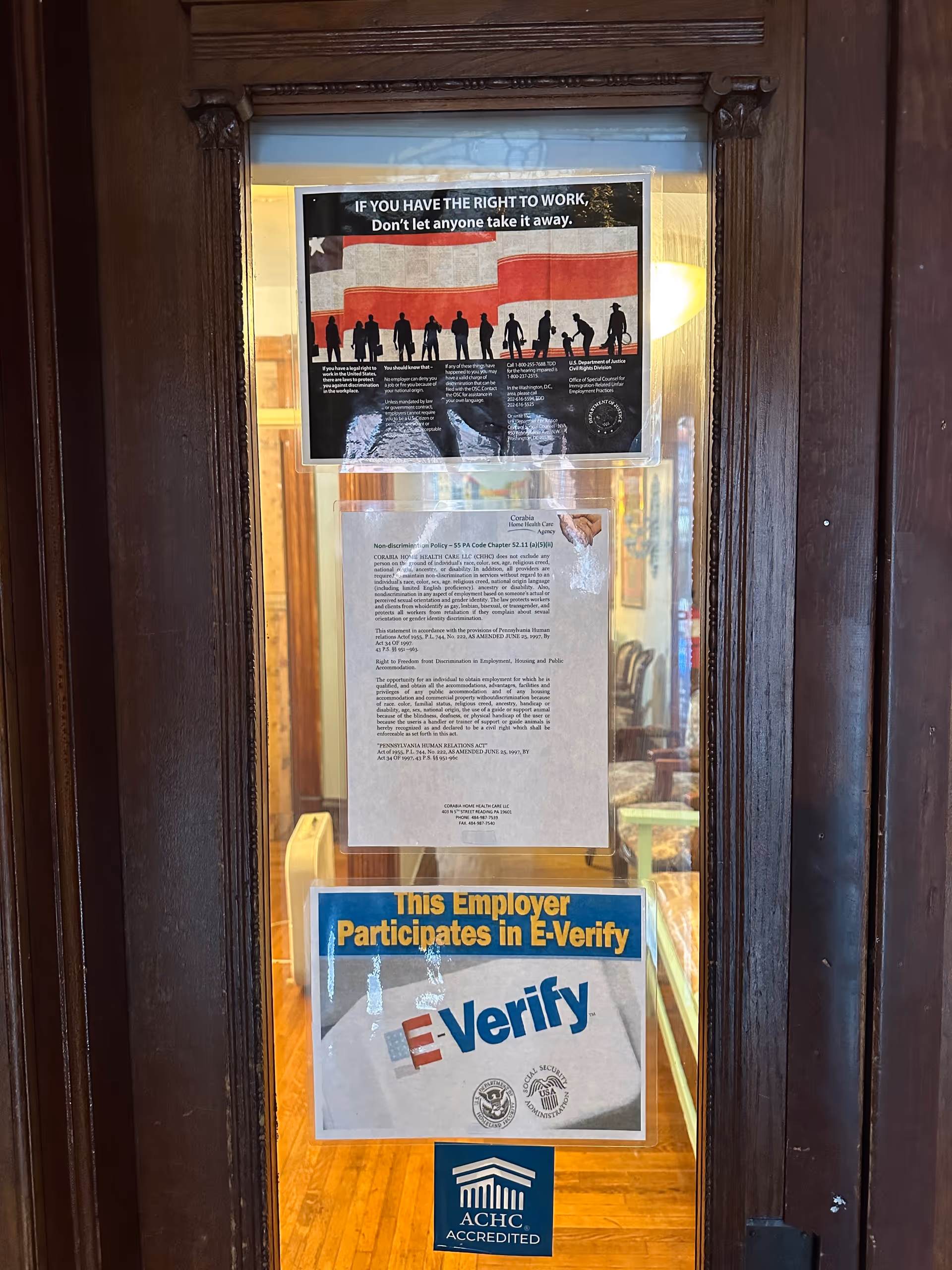A wooden framed glass door with multiple posted notices, including E-Verify and accreditation signs at entrance.