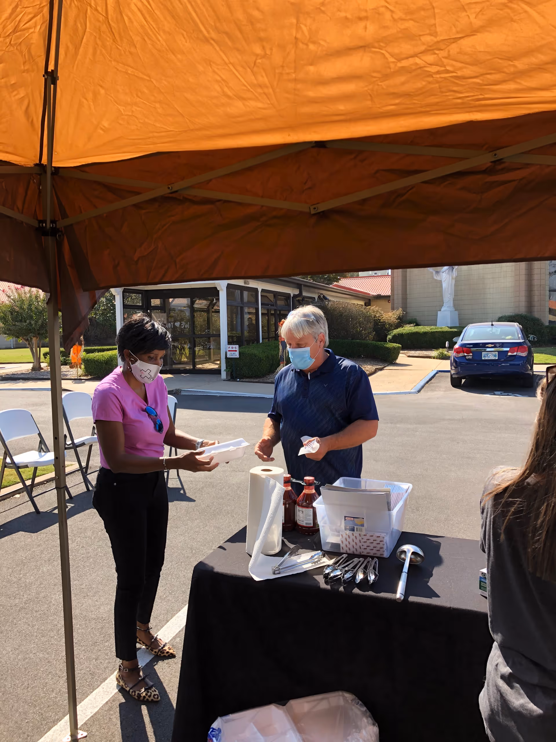 Two masked people stand at an outdoor table under an orange canopy.
