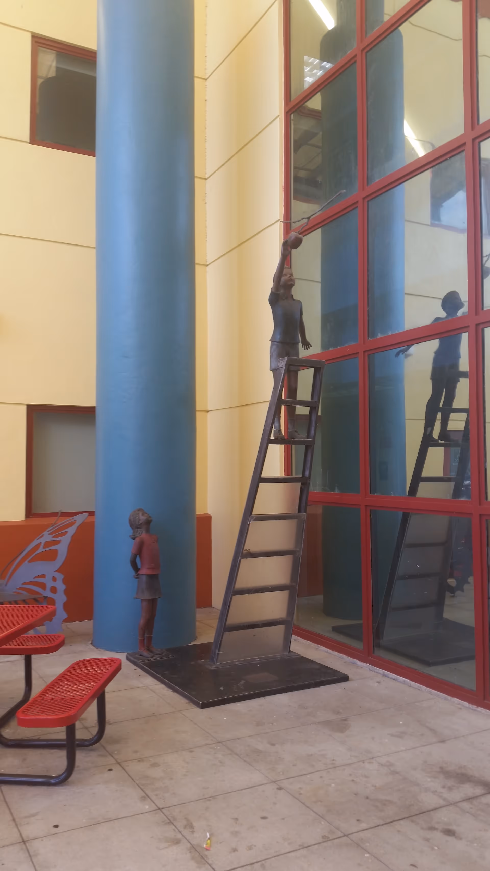 Colorful indoor atrium with blue columns, red-framed glass walls, and statues on ladders.