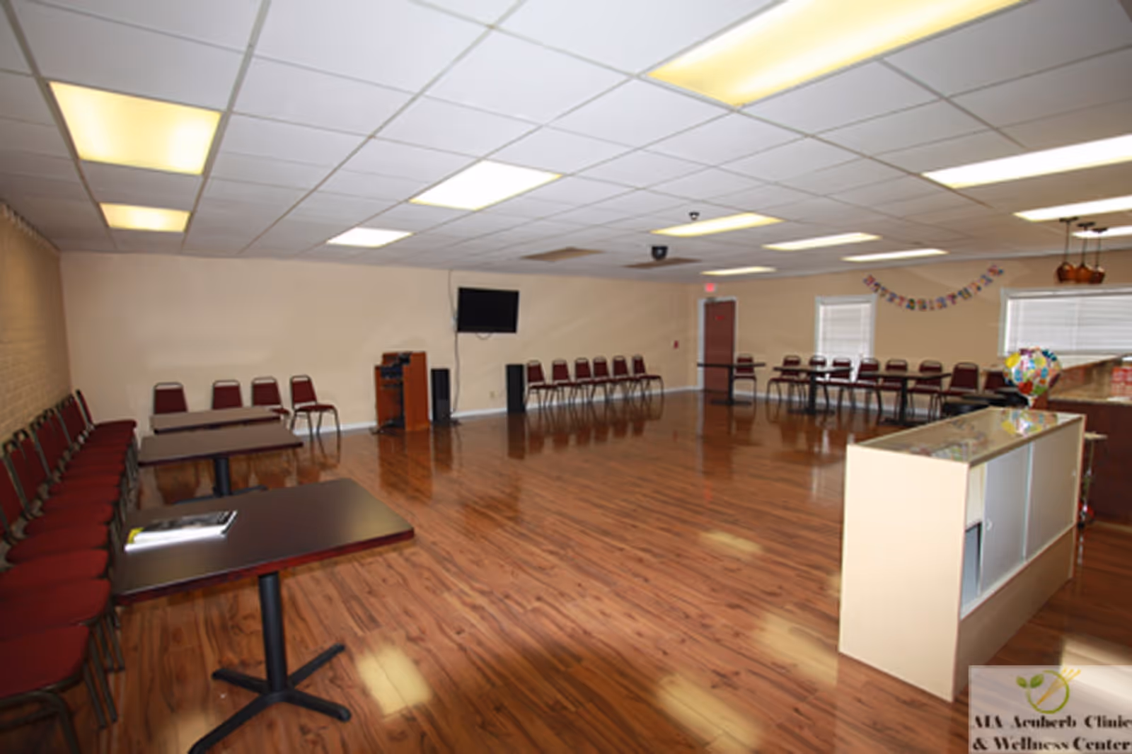 Spacious multipurpose interior room with wooden floor, rows of chairs along walls, and a reception counter.