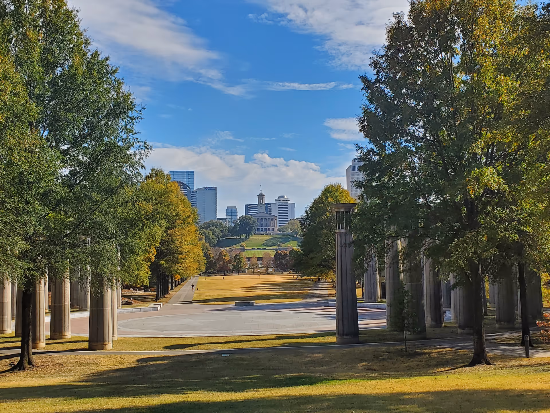 Park-like avenue with tall columns, trees, and distant city skyline under blue sky