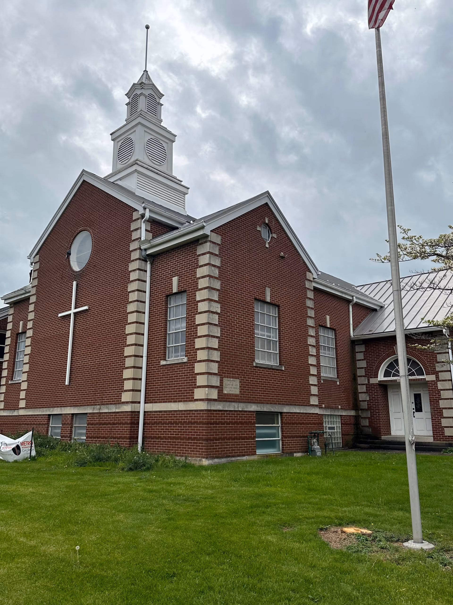 Brick church-style building with a white steeple, cross on the wall, flagpole, and green lawn.