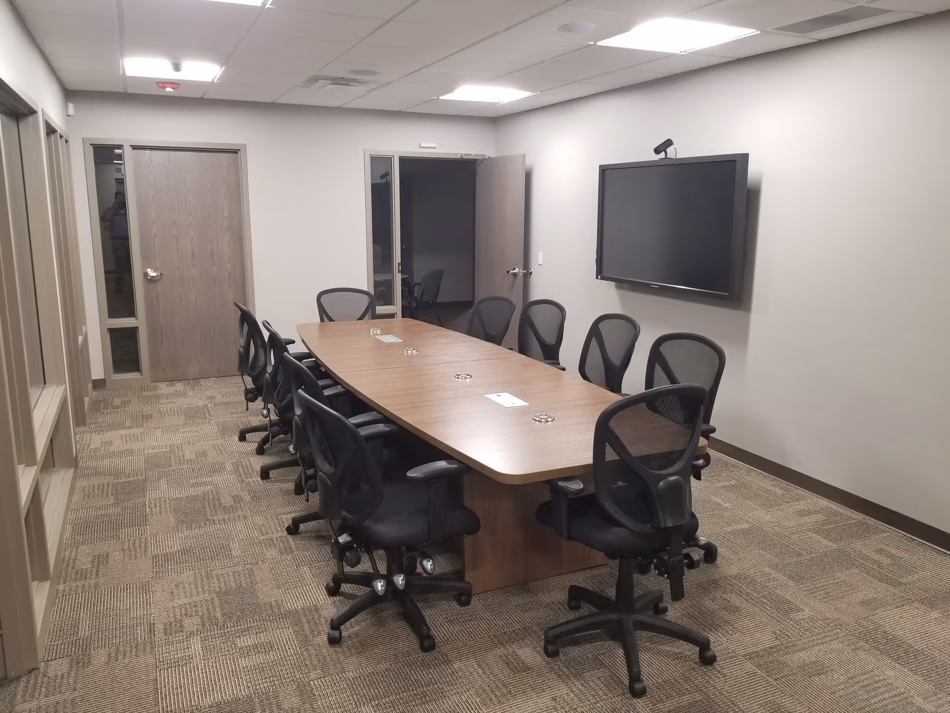 A long conference table with chairs in a neutral, office-like meeting room with a wall screen.