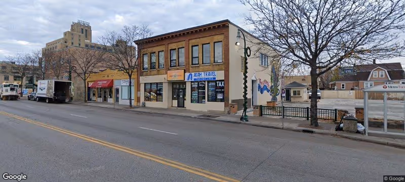 Exterior street view of a row of storefronts and trees along a wide road.