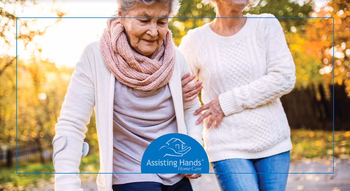 Two elderly women walk outdoors on a sunny autumn day; Assisting Hands Home Care logo visible.