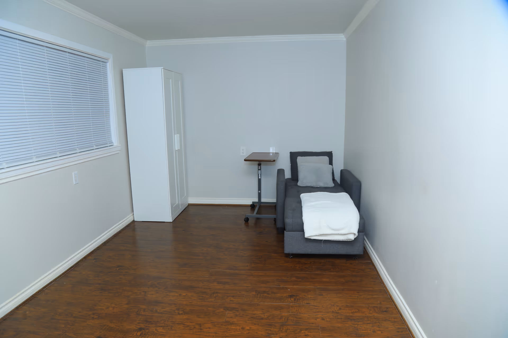 Small, sparsely furnished room with a gray sofa, wardrobe, small table, and wooden floor.