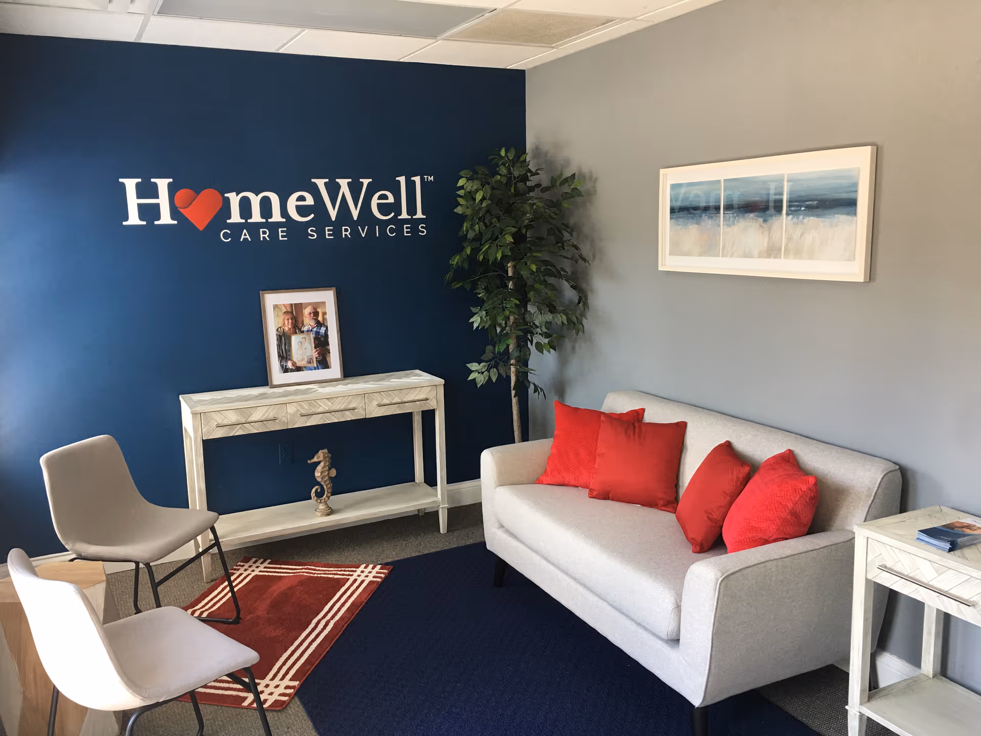 Blue-walled reception lounge with HomeWell Care Services logo, sofa, chairs, plant, and artwork.