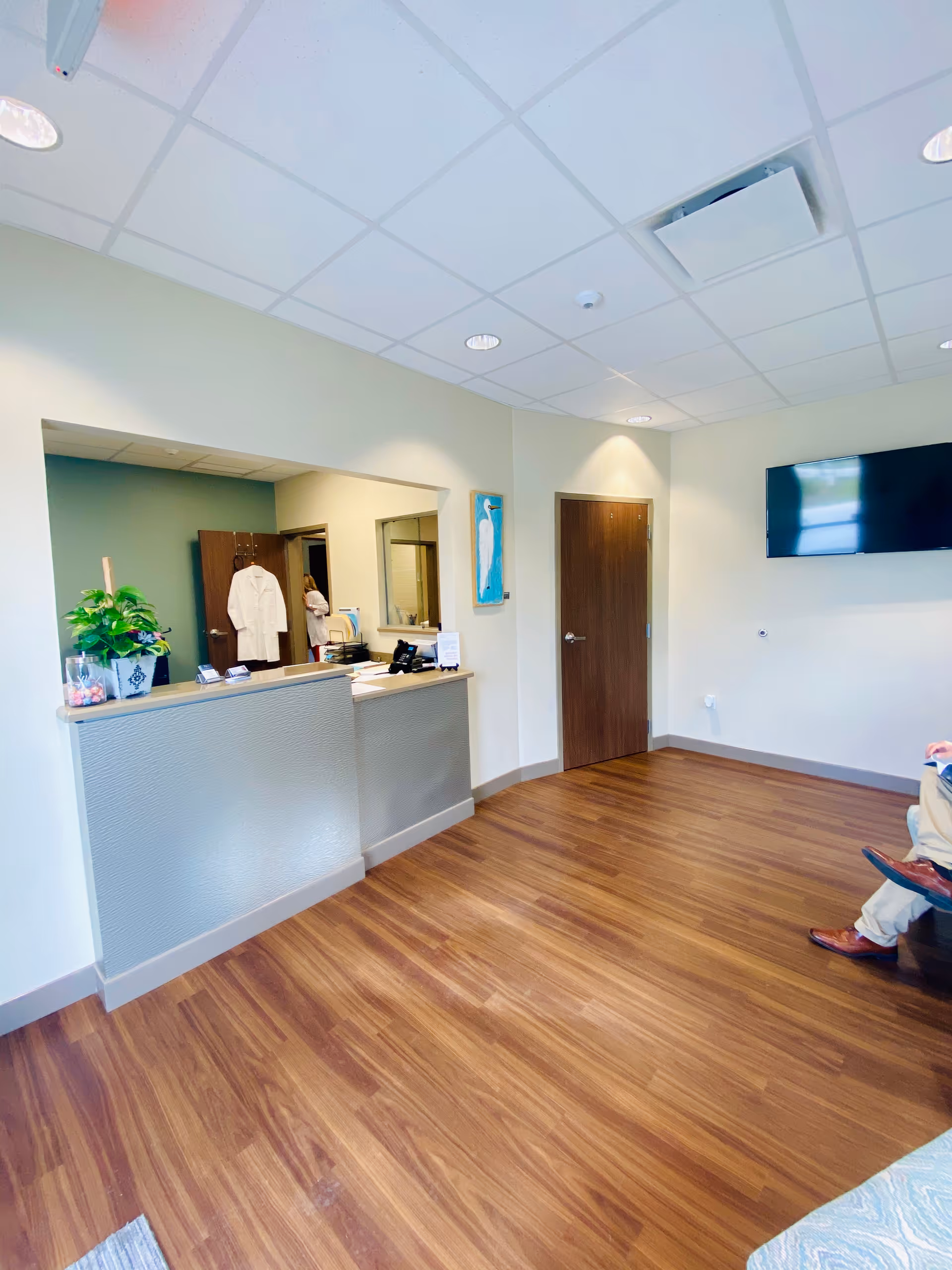 Reception desk in a bright lobby with wood floors, plants, and a wall-mounted TV.