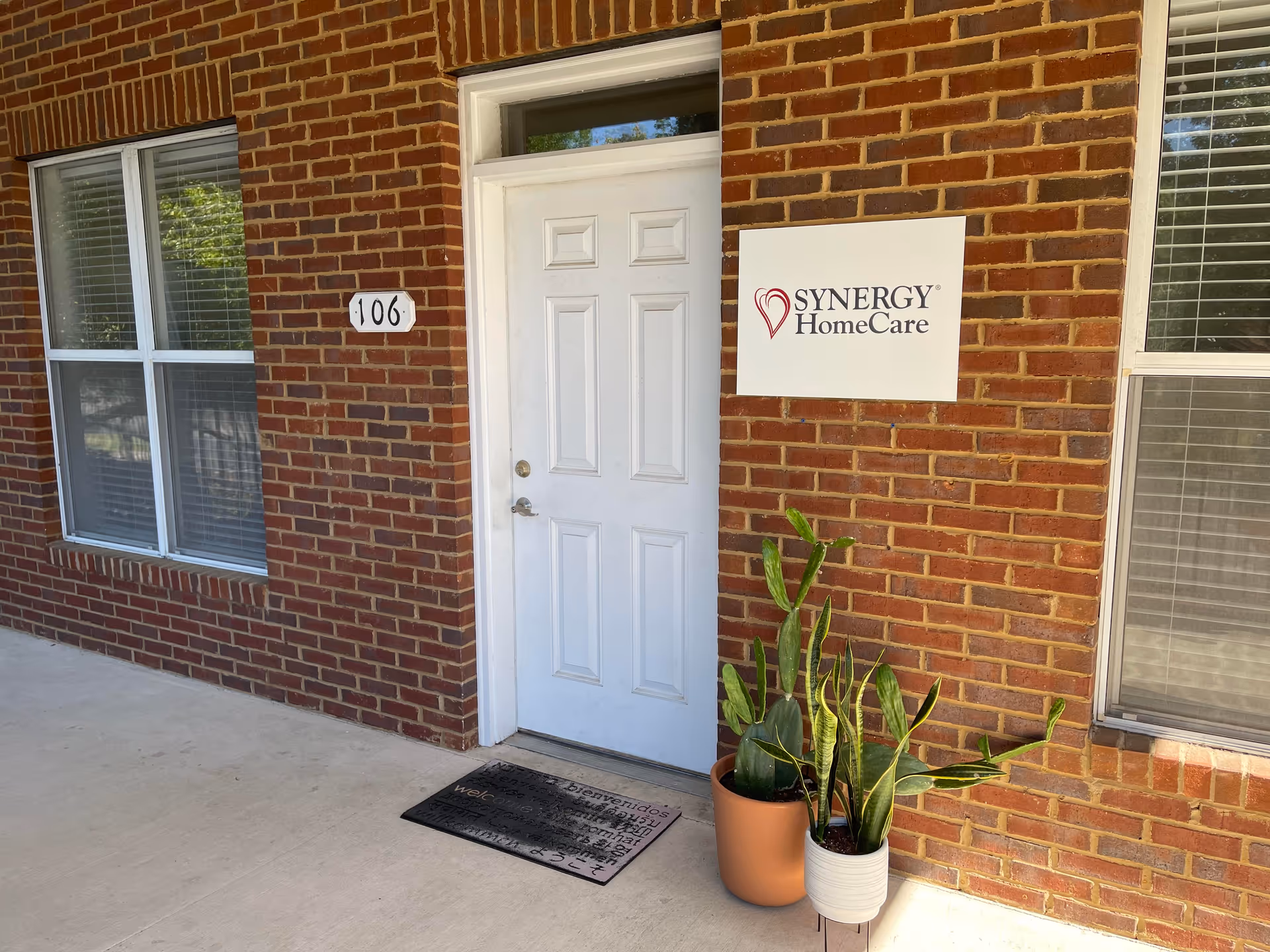 Exterior brick facade with Synergy HomeCare sign beside a white front door and potted plants.