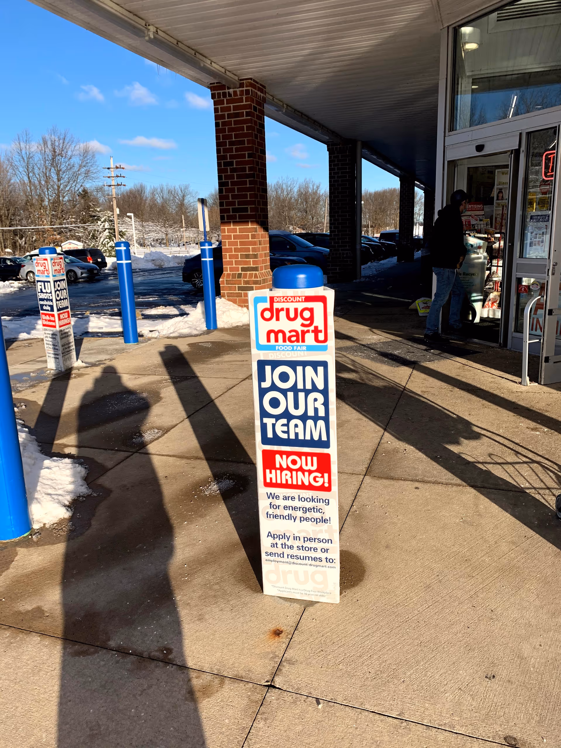 Exterior view of Discount Drug Mart with a tall 'Join Our Team' sign outside.