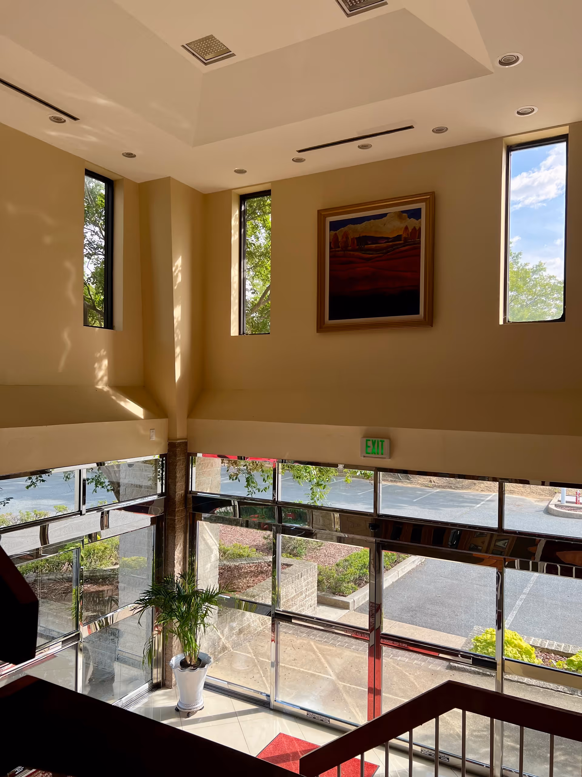 Bright interior atrium with tall windows, framed painting, potted plant, and glass entry doors.