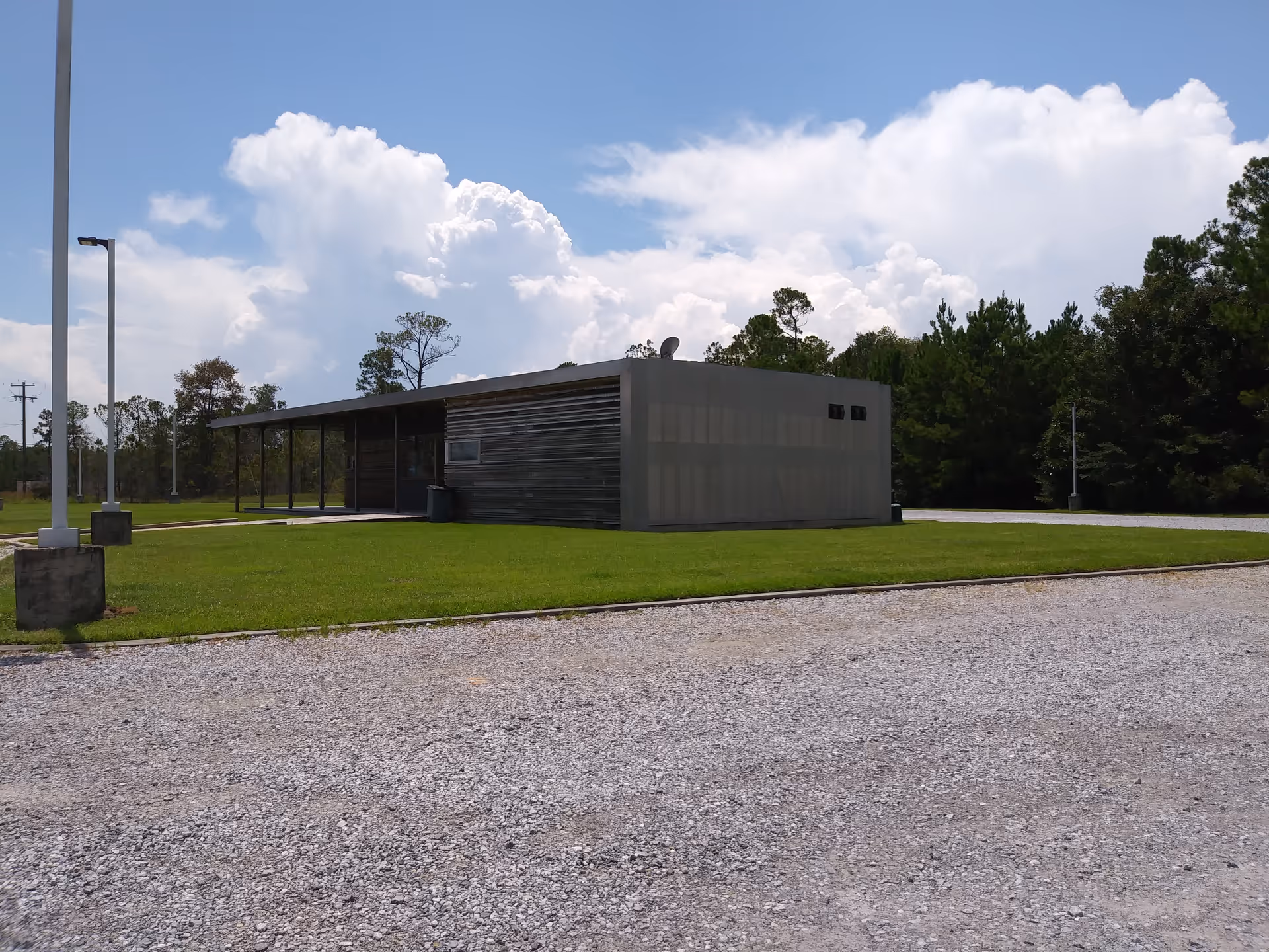 A single-story modern facility building sits on a grassy lot with gravel foreground and trees.