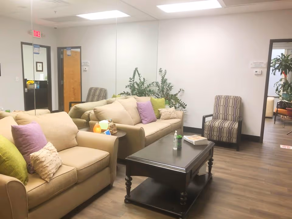 A cozy living room lounge with beige sofas, colorful pillows, a dark coffee table, and plants.