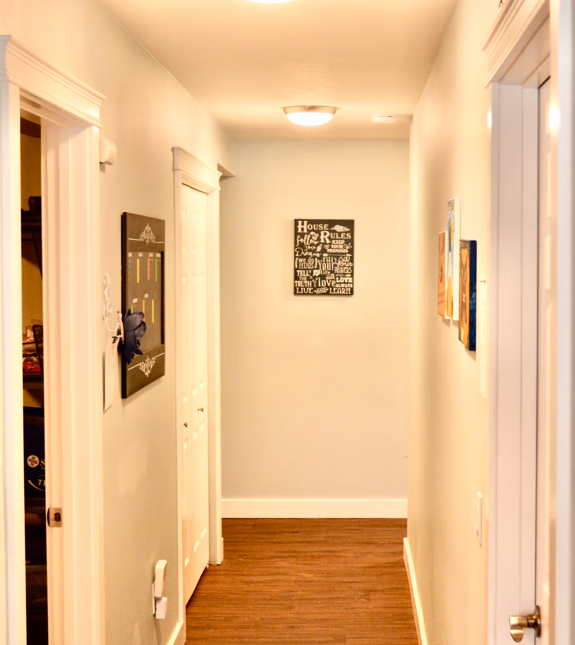 Bright hallway in a senior living facility with doors, wall art, and wood flooring.