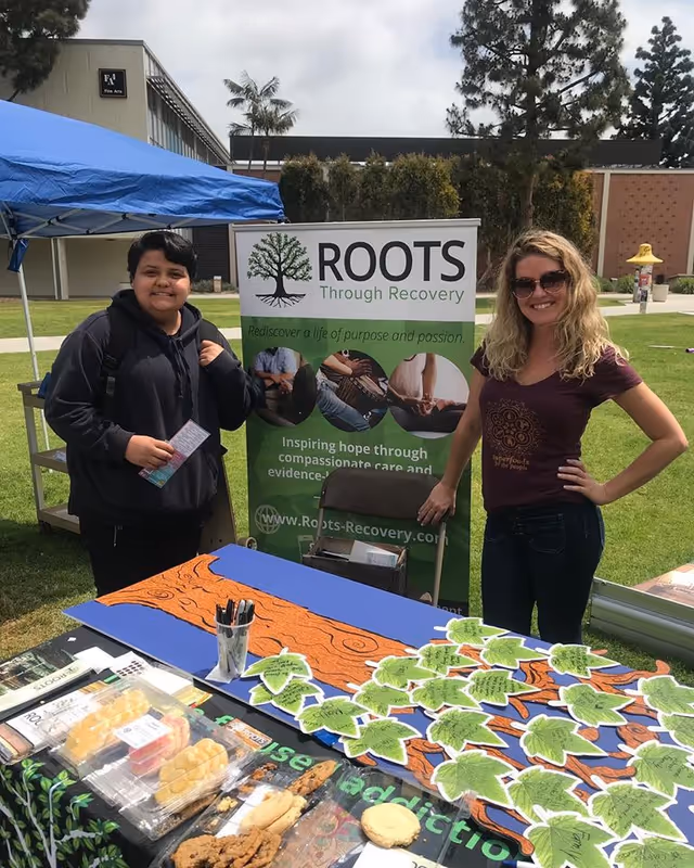 Two people stand beside a Roots Through Recovery booth at an outdoor event.