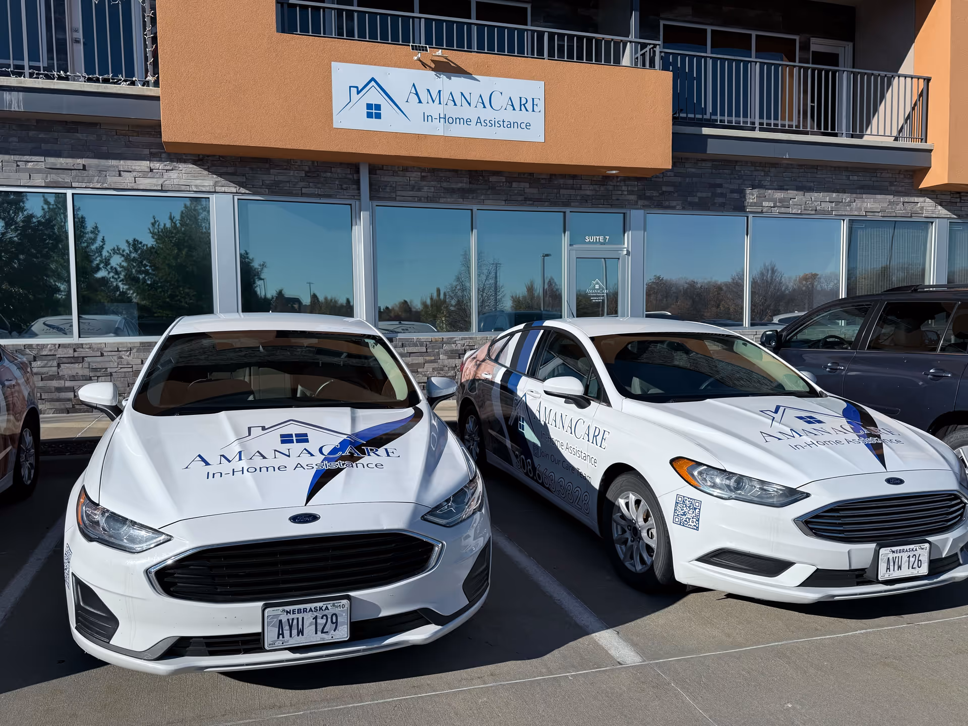 Exterior view of AmanaCare facility with sign, storefront windows, and two parked cars.
