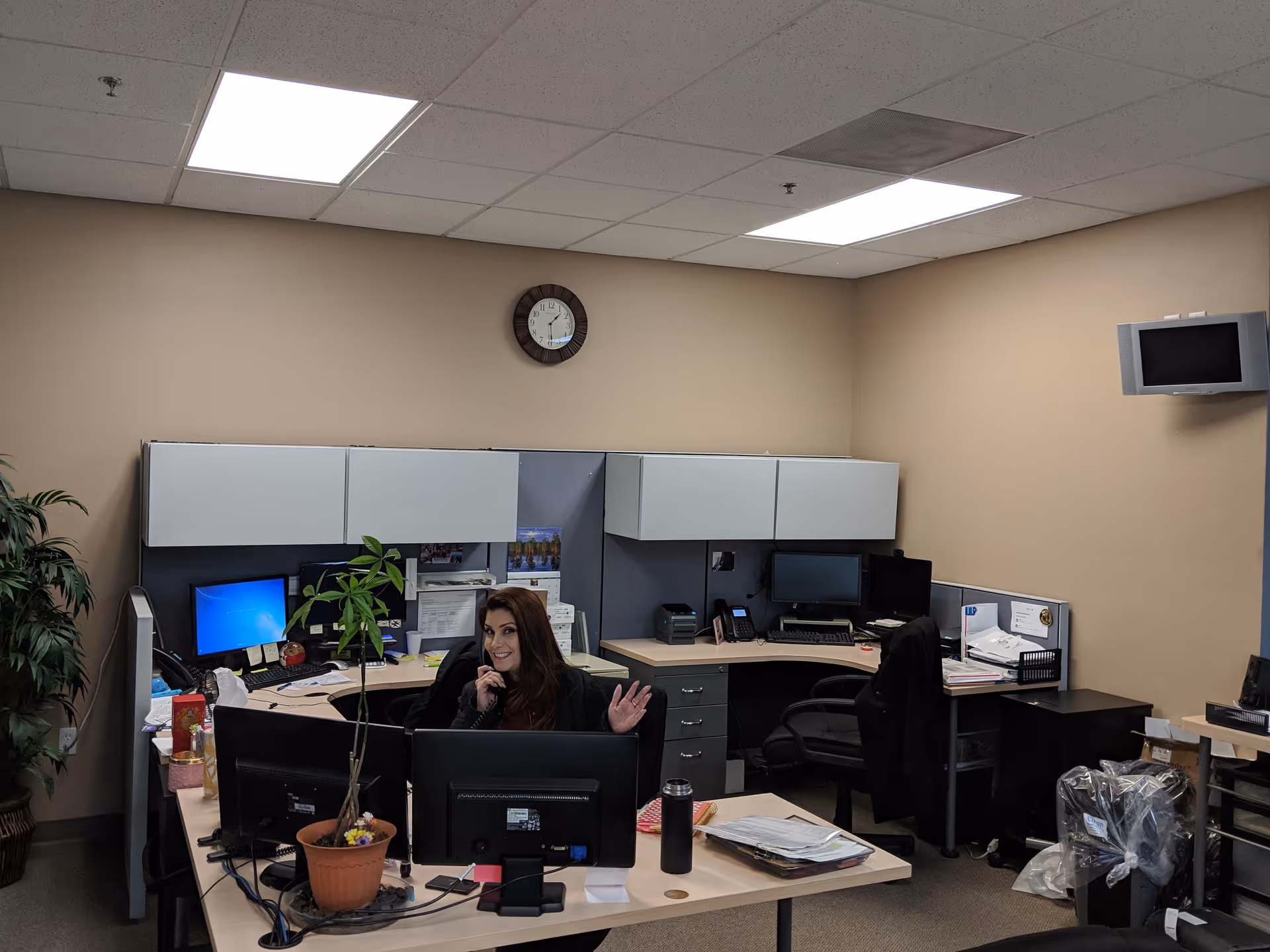 Office cubicles with a woman at a desk, computer monitors, and a clock on beige walls