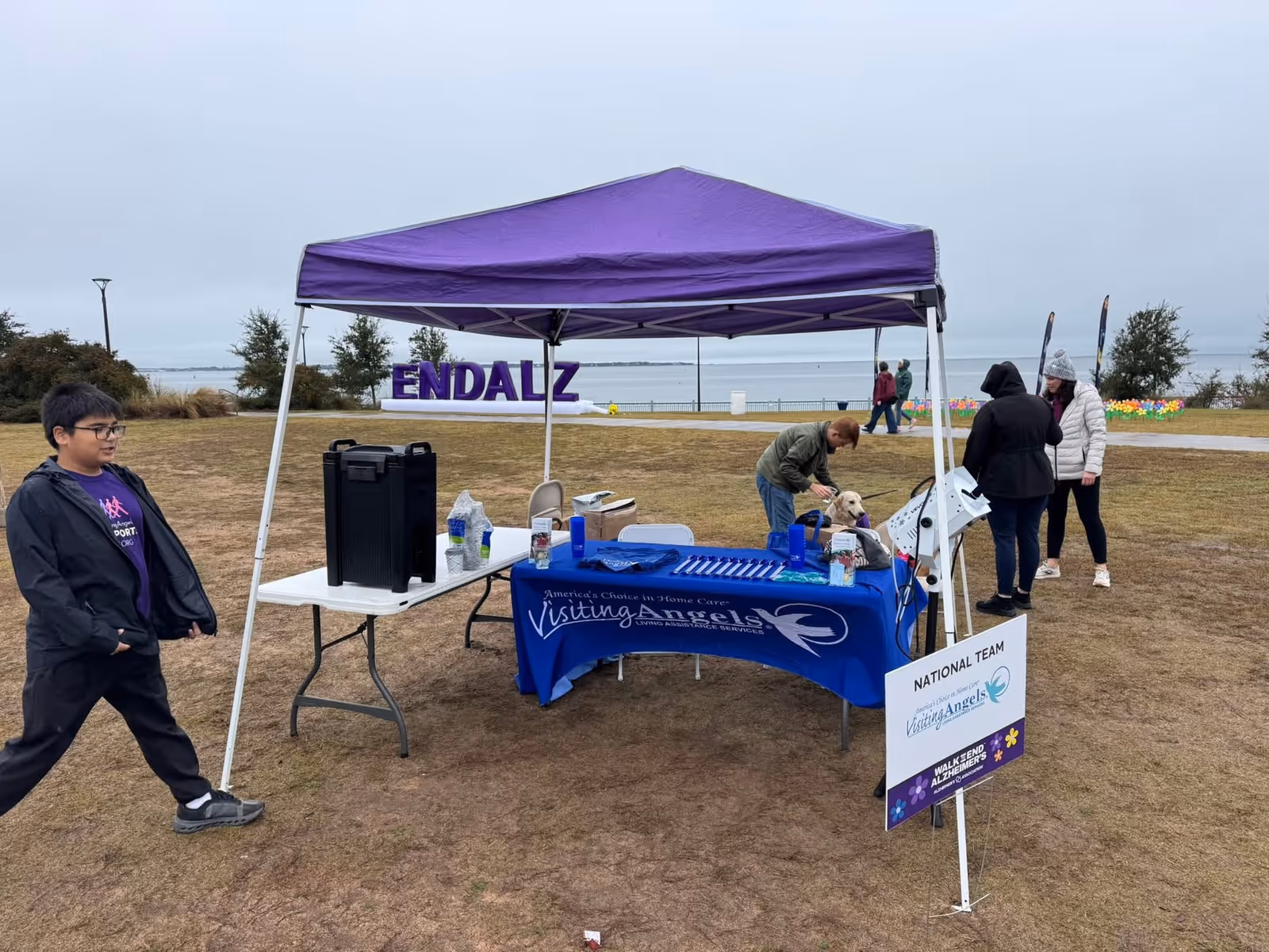 Purple canopy over a Visiting Angels booth at an outdoor event near water.