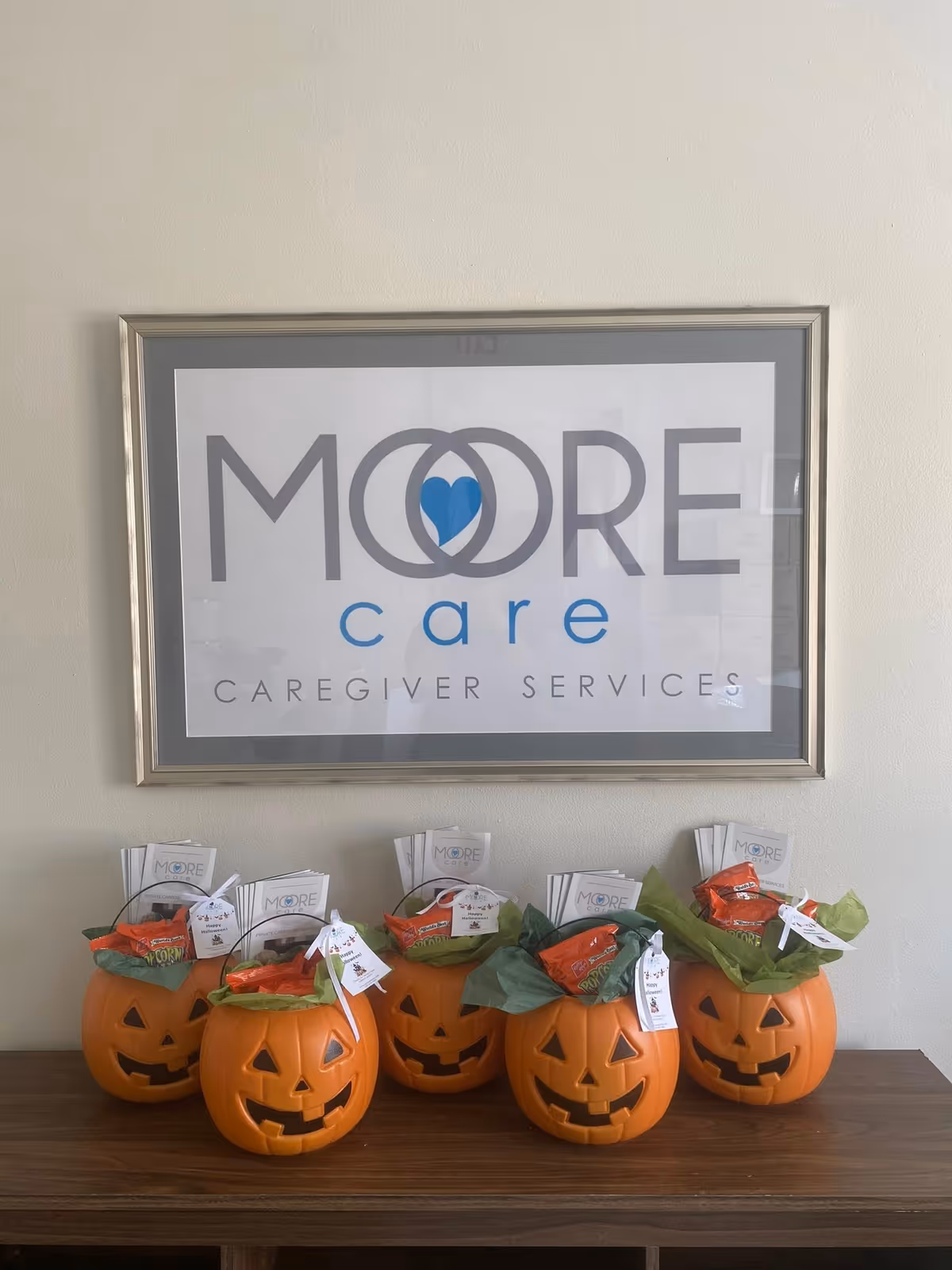 Framed Moore Care logo on wall above a table with jack-o'-lantern pumpkins.