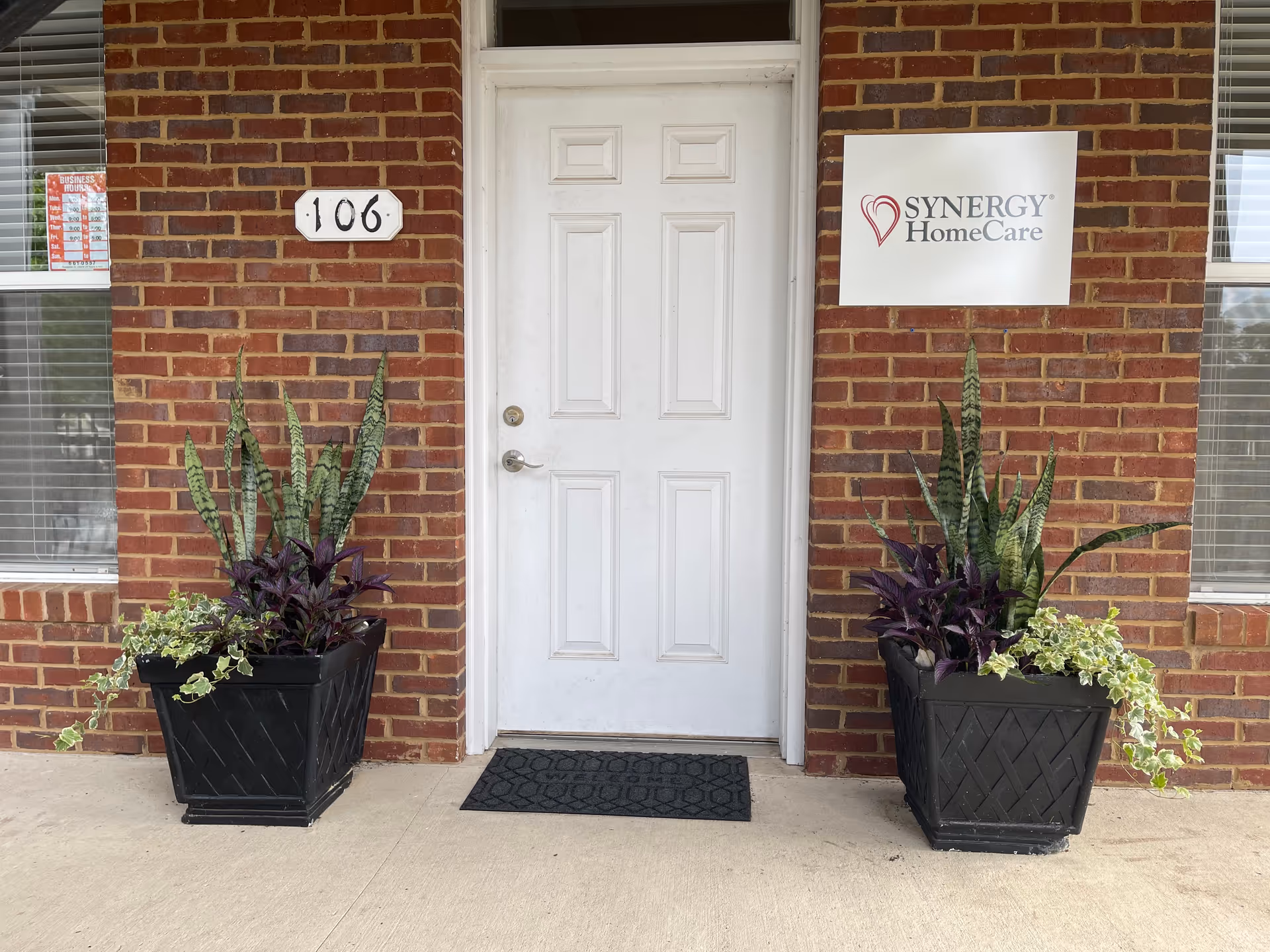 Brick building entrance with white door, two planters, and Synergy HomeCare sign.