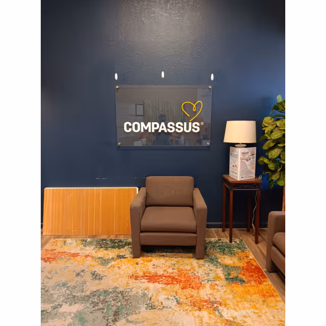Dark blue wall with Compassus sign, brown armchair, side table, lamp, plant, and colorful rug.