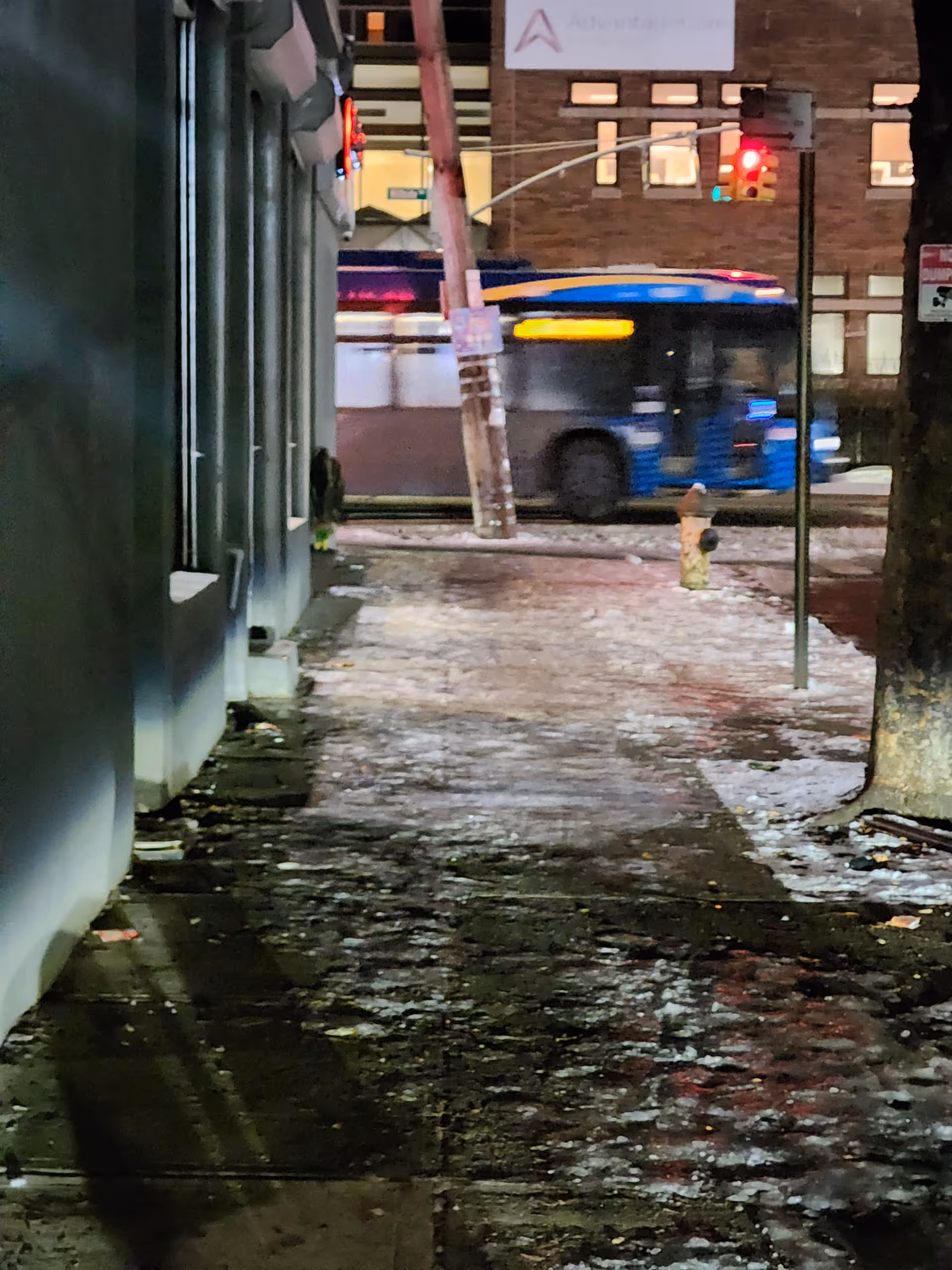 Nighttime exterior scene of a city sidewalk beside a building with a moving bus.