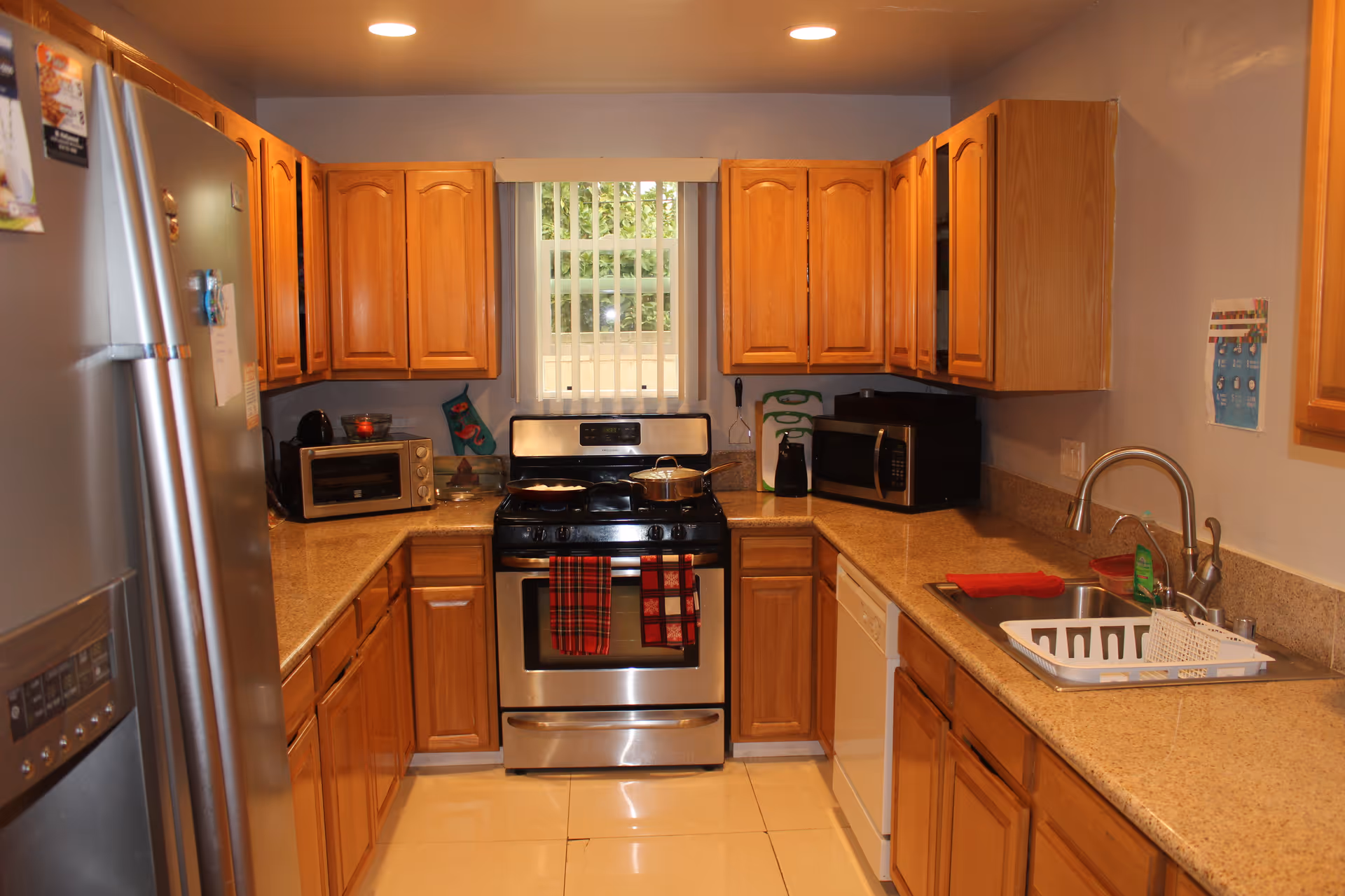 Compact kitchen with wooden cabinets, stainless stove, fridge, microwave, and sink under a window.