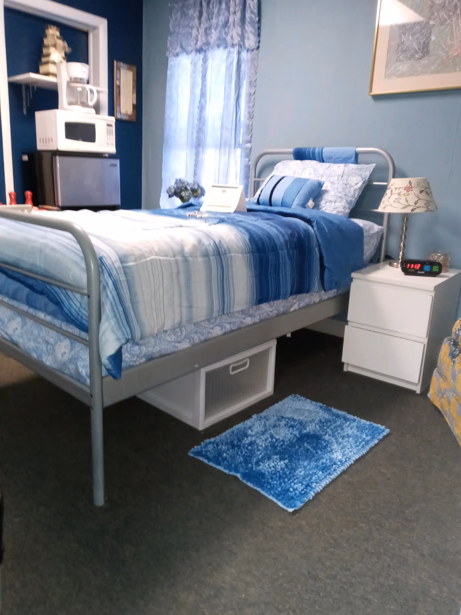 Blue-themed bedroom with metal bed, nightstand, lamp, window, and small appliances.