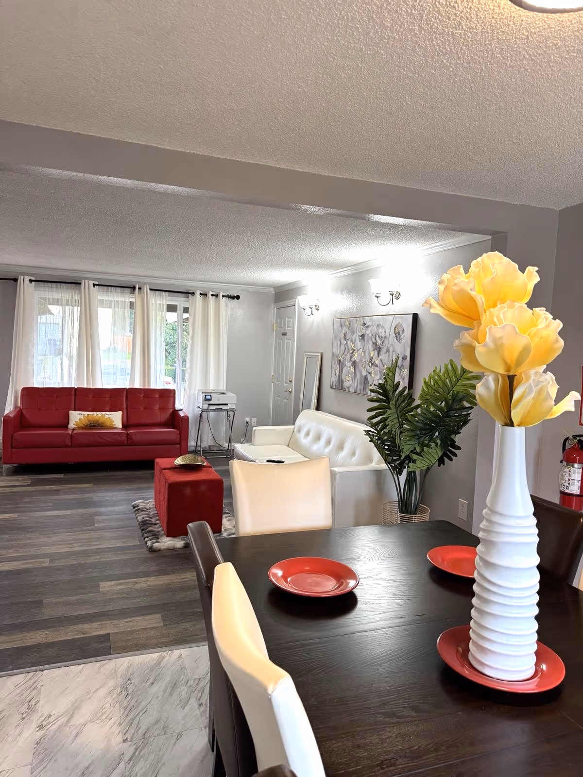 Open living and dining area with red sofa, white seating, plants, and a tall vase centerpiece.