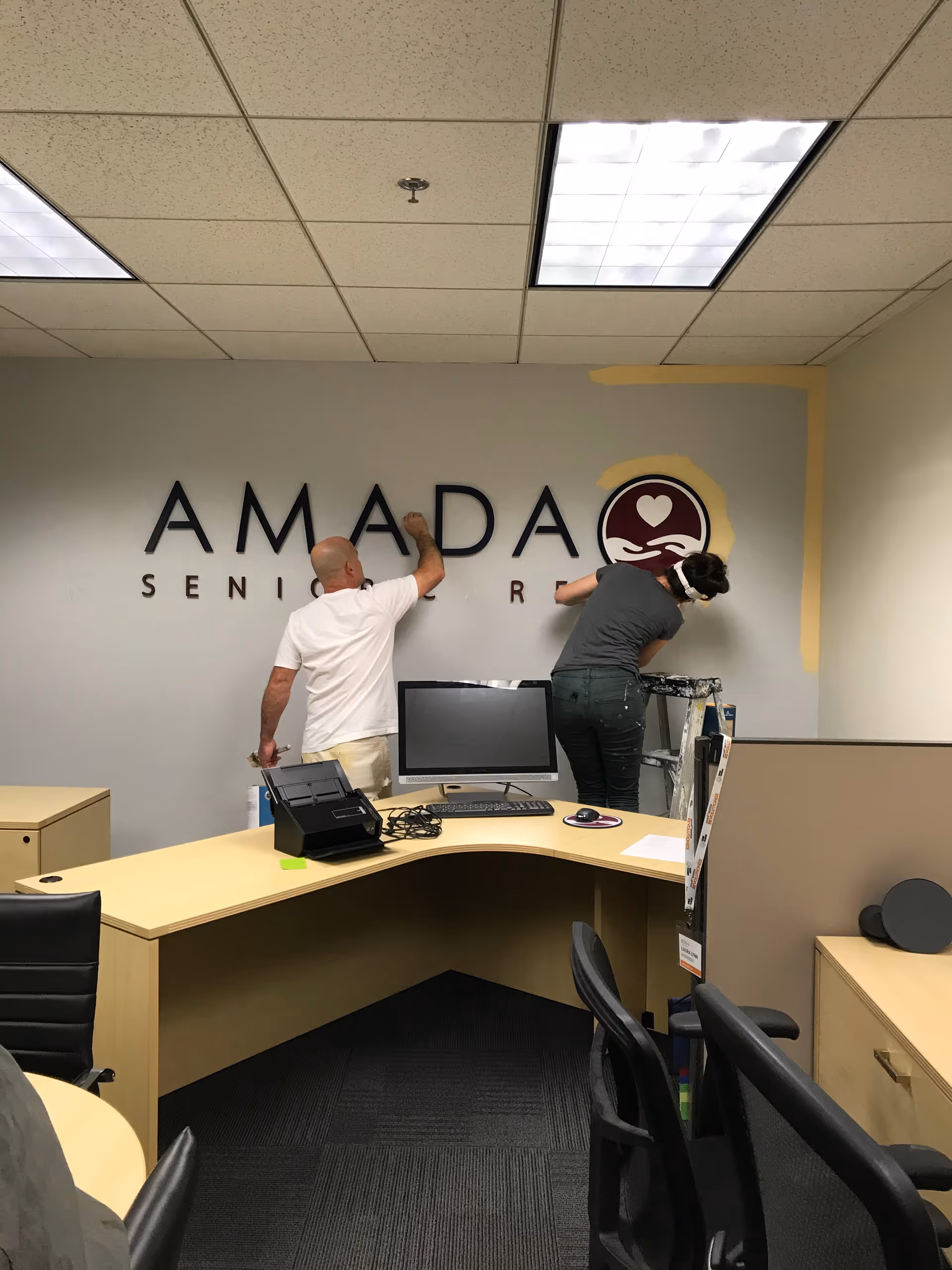 Two staff members install an Amada Senior Care logo on a wall in a reception area.