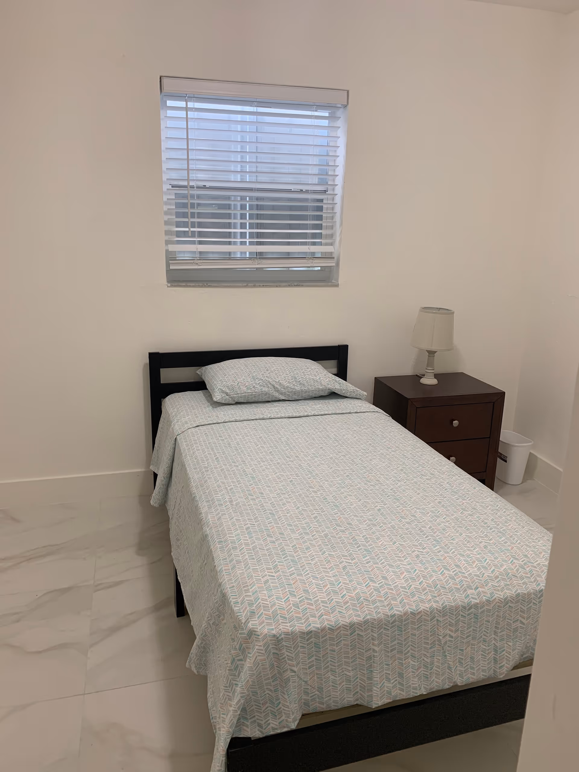 Small bedroom with single bed, nightstand, lamp, and window blinds.
