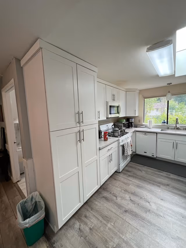 White modern kitchen with tall cabinets, stove, microwave, dishwasher, and sink near a window.