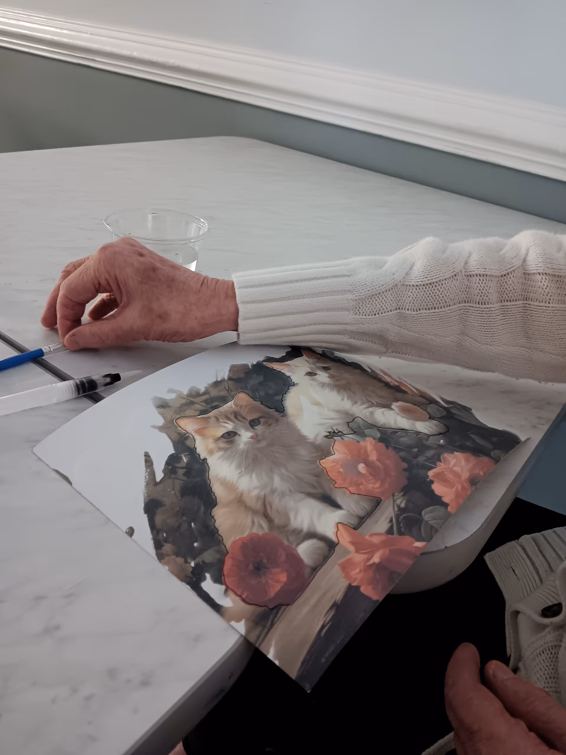 An elderly hand rests on a marble table beside a painting and a cup.