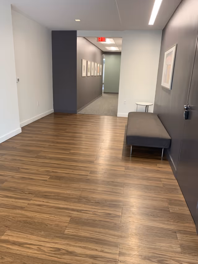 Empty hallway in a modern facility with wood flooring, a gray bench, and framed art.