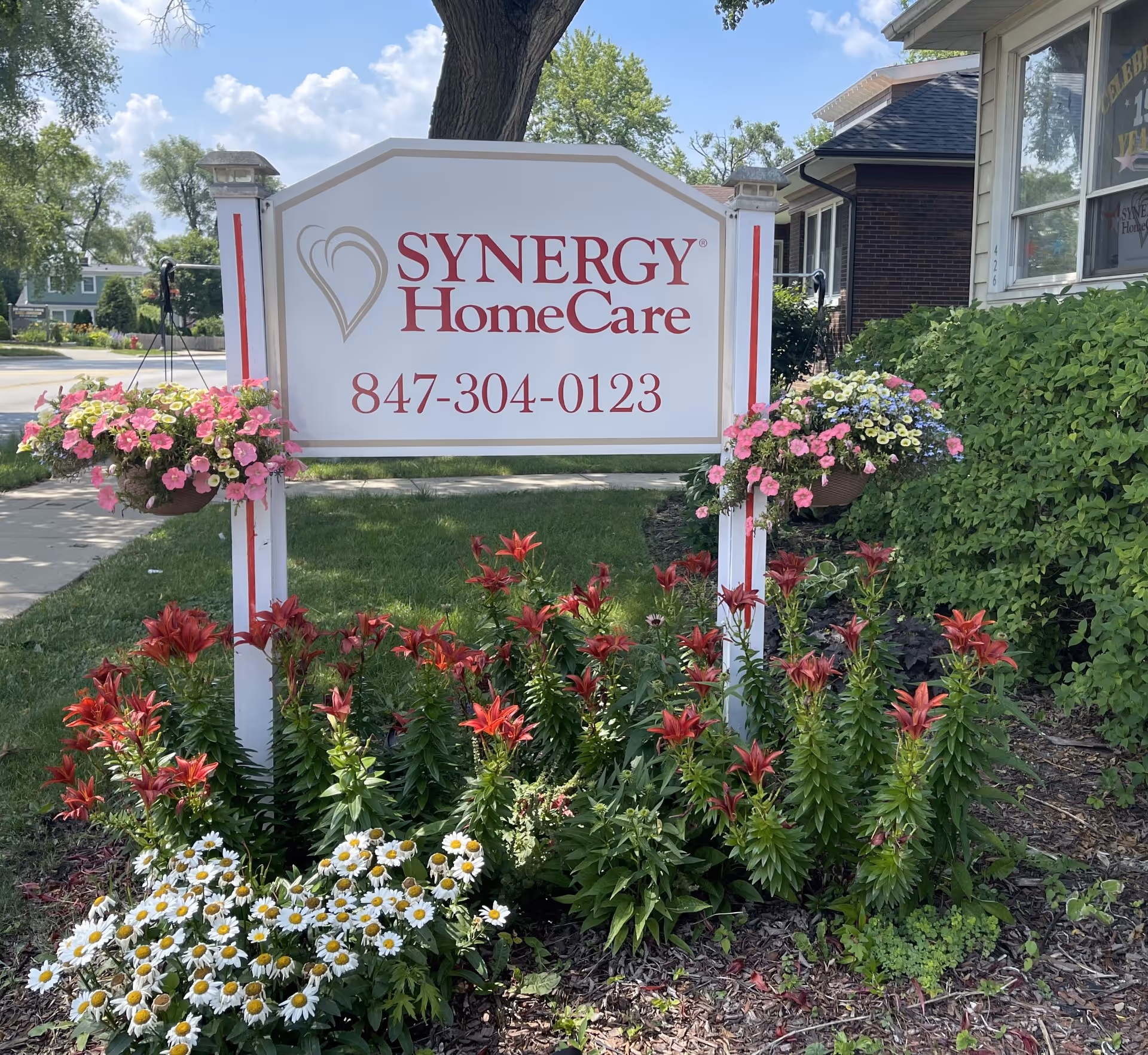 SYNERGY HomeCare sign outdoors with colorful flowers and greenery.
