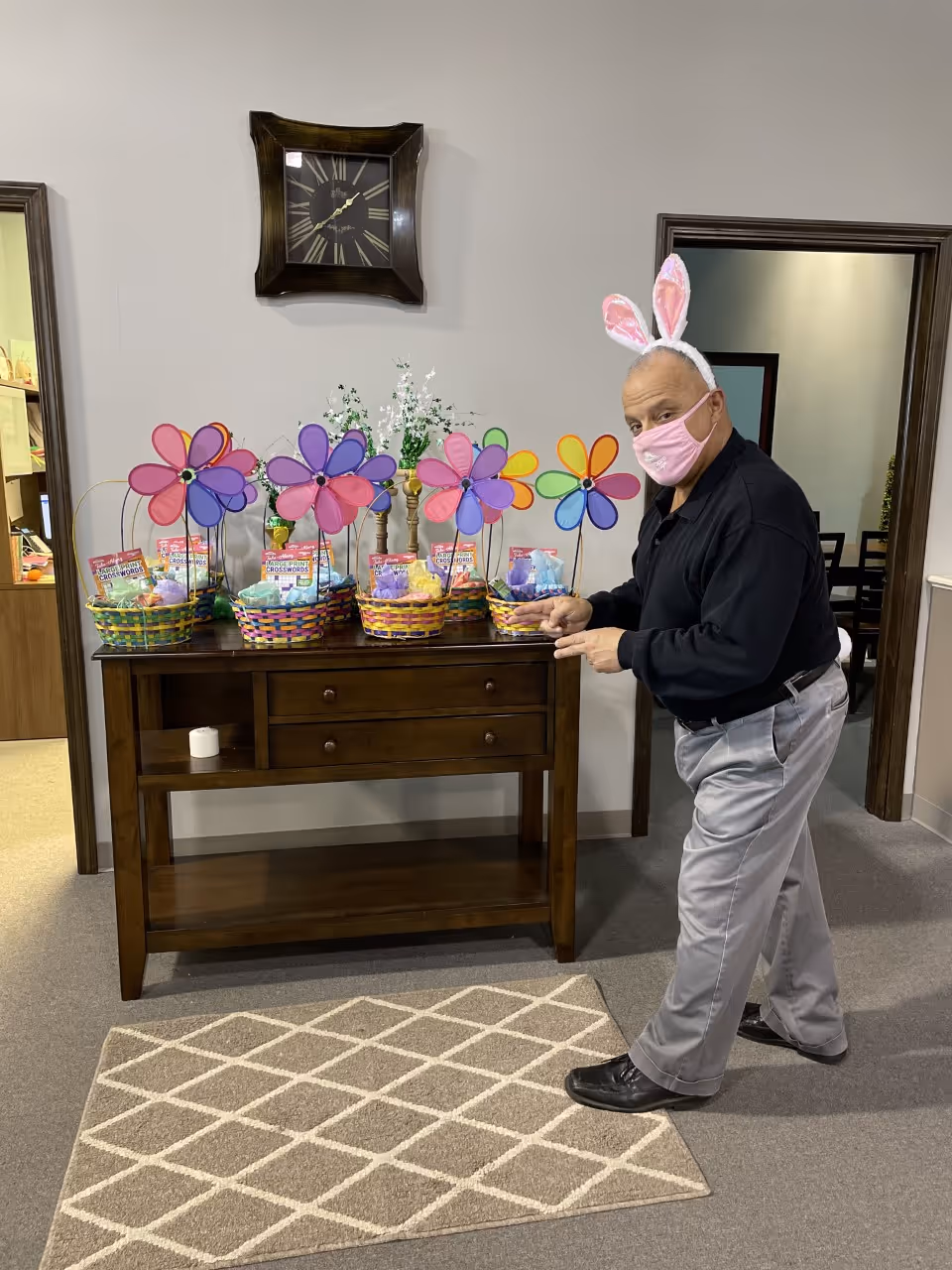 Staff member wearing bunny ears and mask arranges Easter baskets on a hallway console.