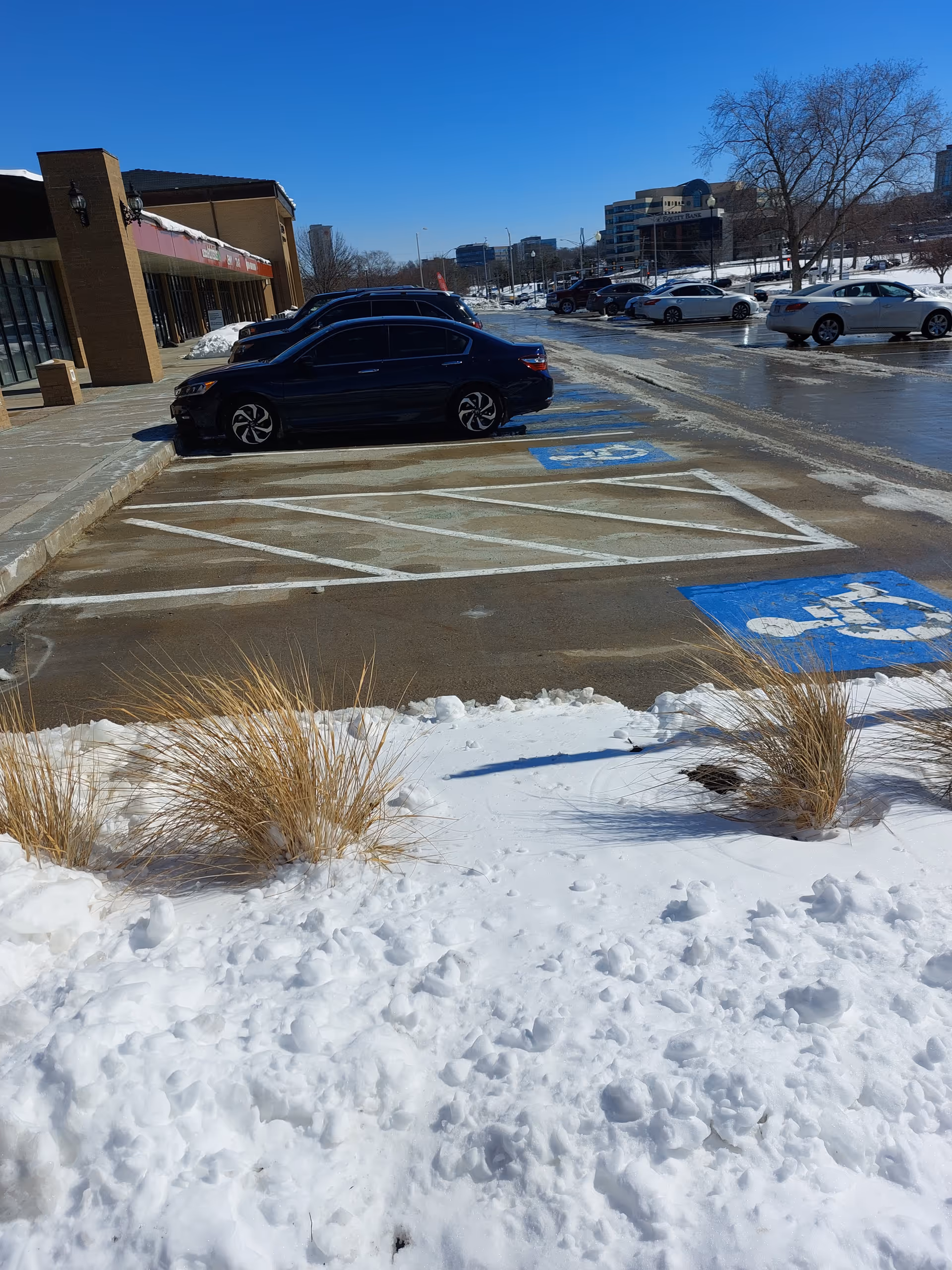 A snowy parking lot outside a strip mall with parked cars and handicap spots.