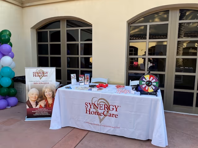 Promotional table with Synergy HomeCare banner outside a beige building, balloons nearby.