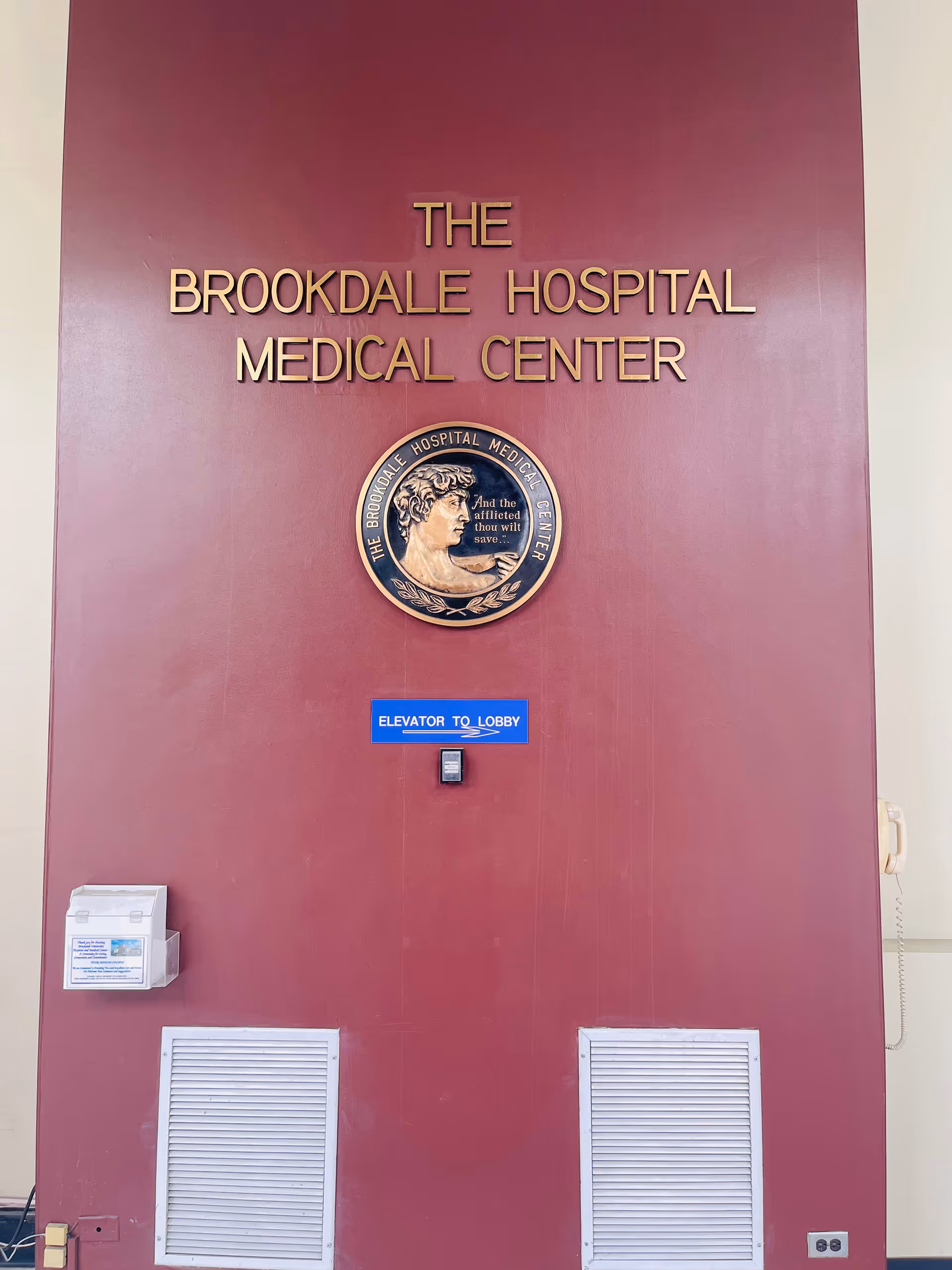 Maroon wall with sign 'The Brookdale Hospital Medical Center', circular emblem, blue Elevator to Lobby sign.