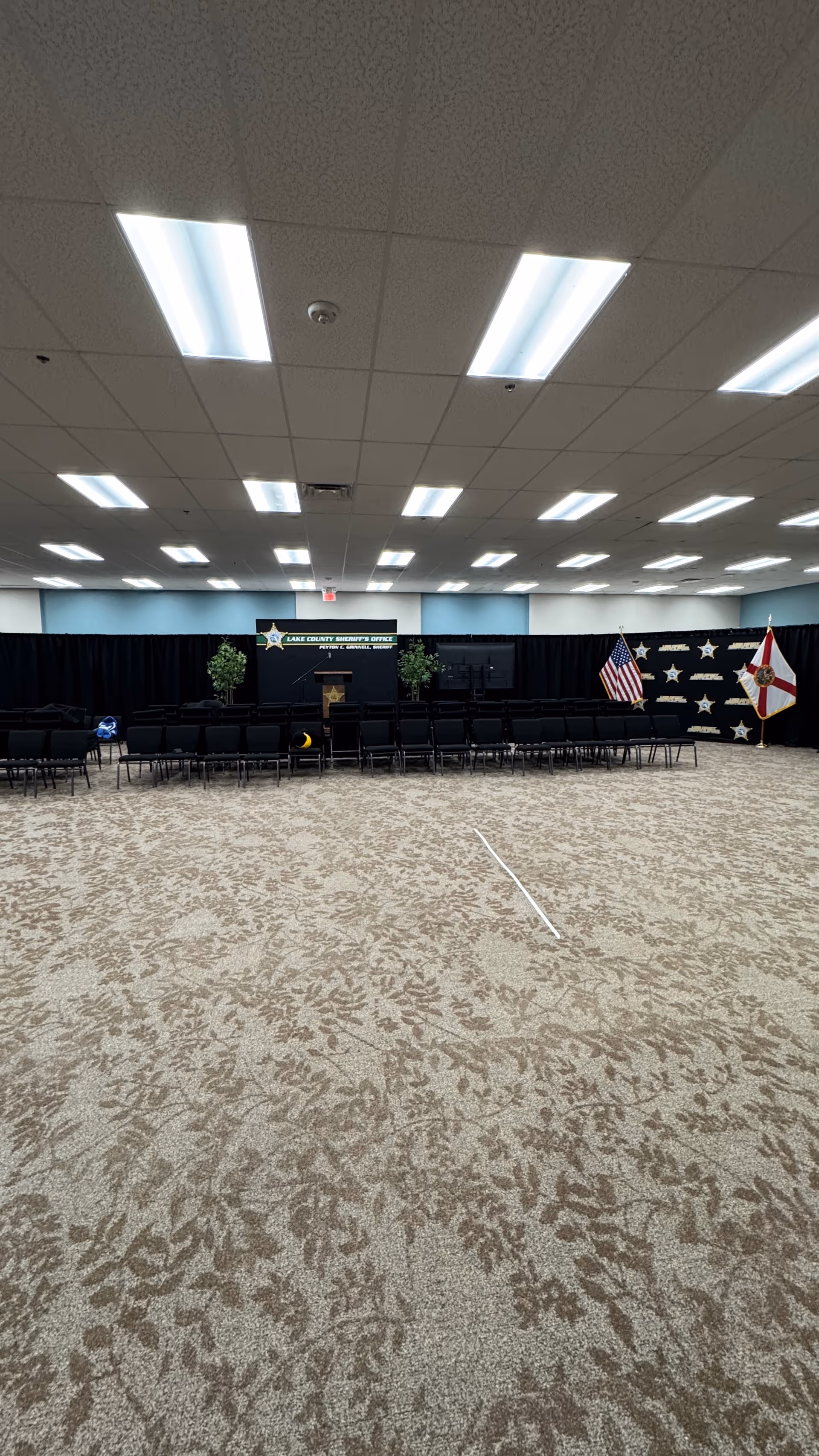 Large indoor hall with a stage, rows of chairs, flags, and patterned carpet.