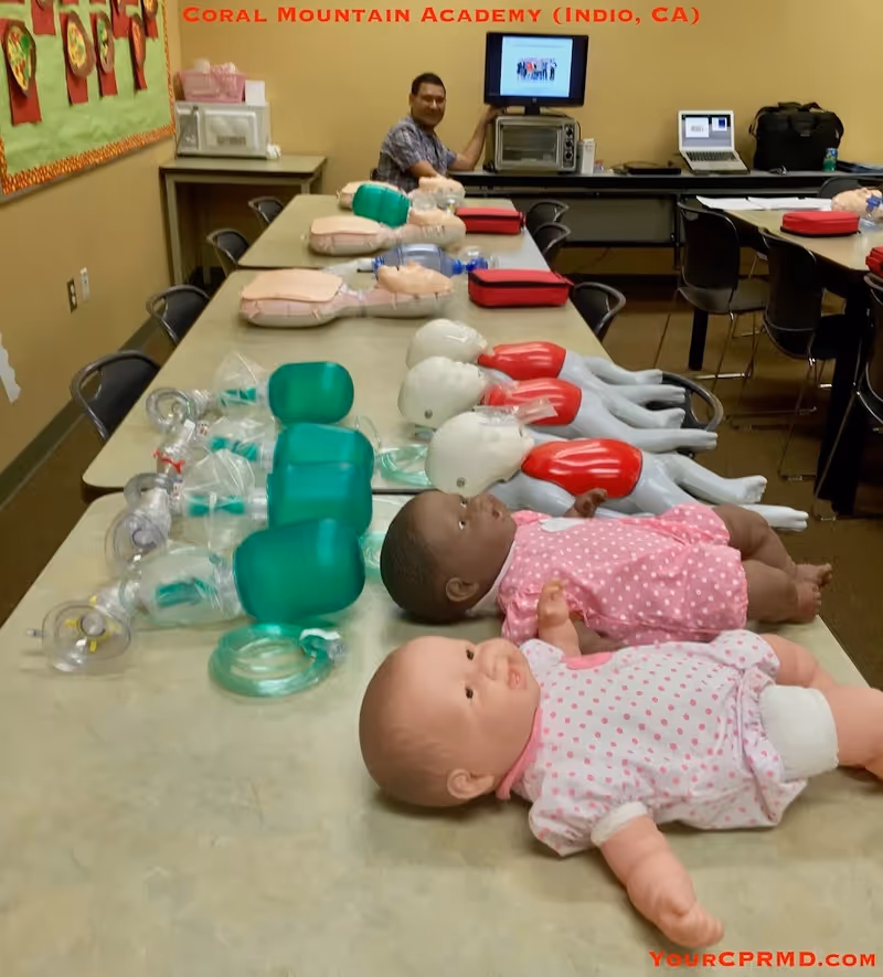 A classroom-style CPR training room with infant and adult manikins and equipment on a long table.