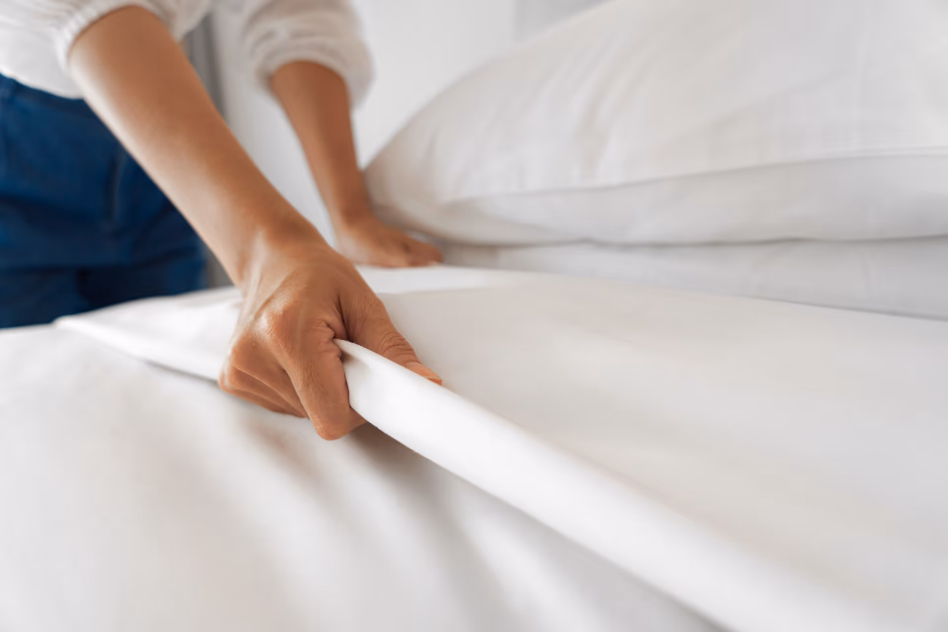 Caregiver tucks white bed sheets while adjusting a bed in a bedroom.