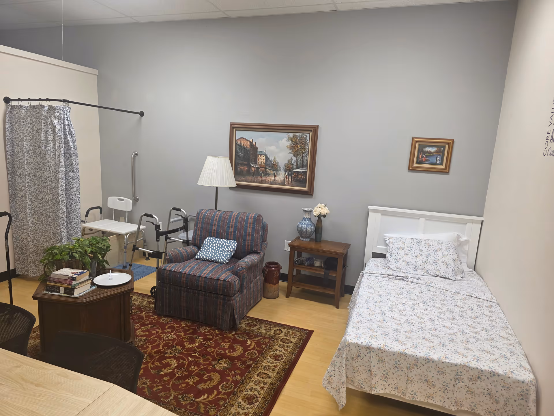 A furnished senior living bedroom with bed, chair, lamp, side table, rug, plant, and mobility aids.