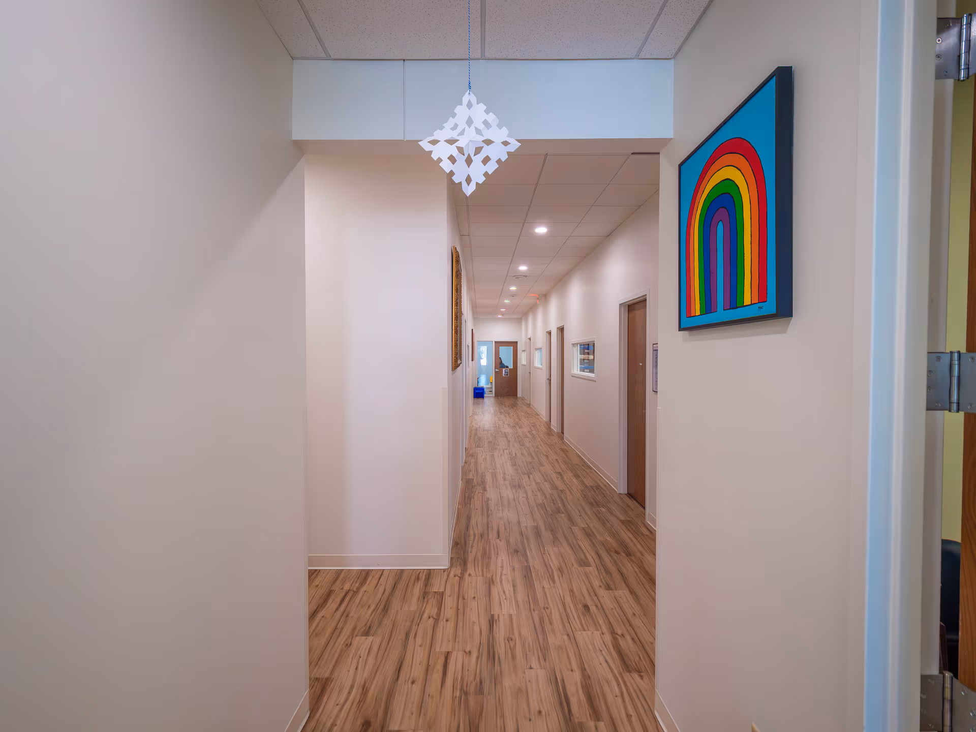 Bright interior hallway of a facility with wooden floors, doors, and rainbow artwork.