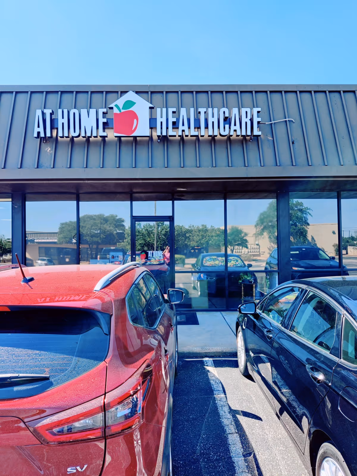 Exterior storefront of At Home Healthcare with glass windows and parked cars.