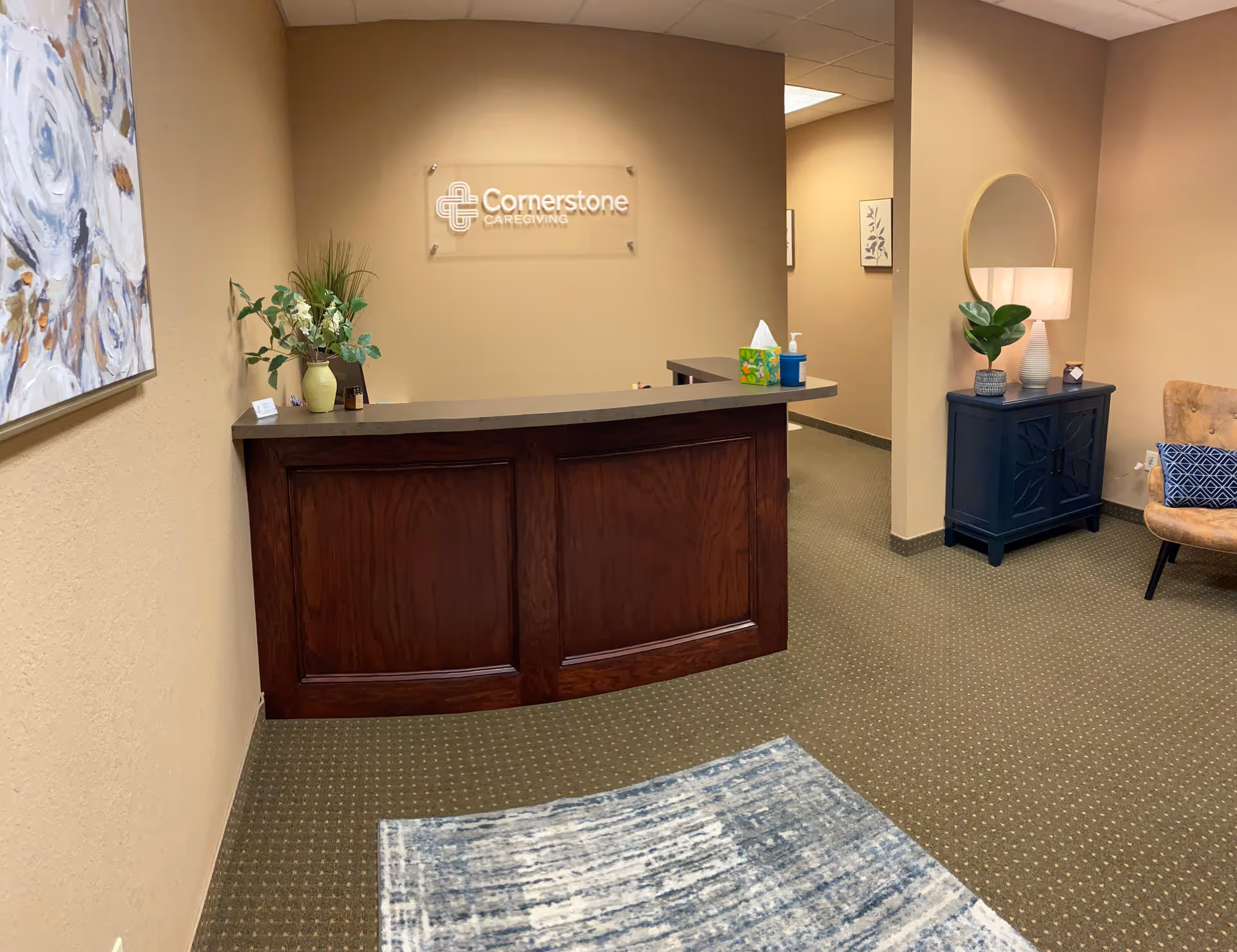 Reception desk in a beige lobby with plants and Cornerstone Caregiving sign on wall.