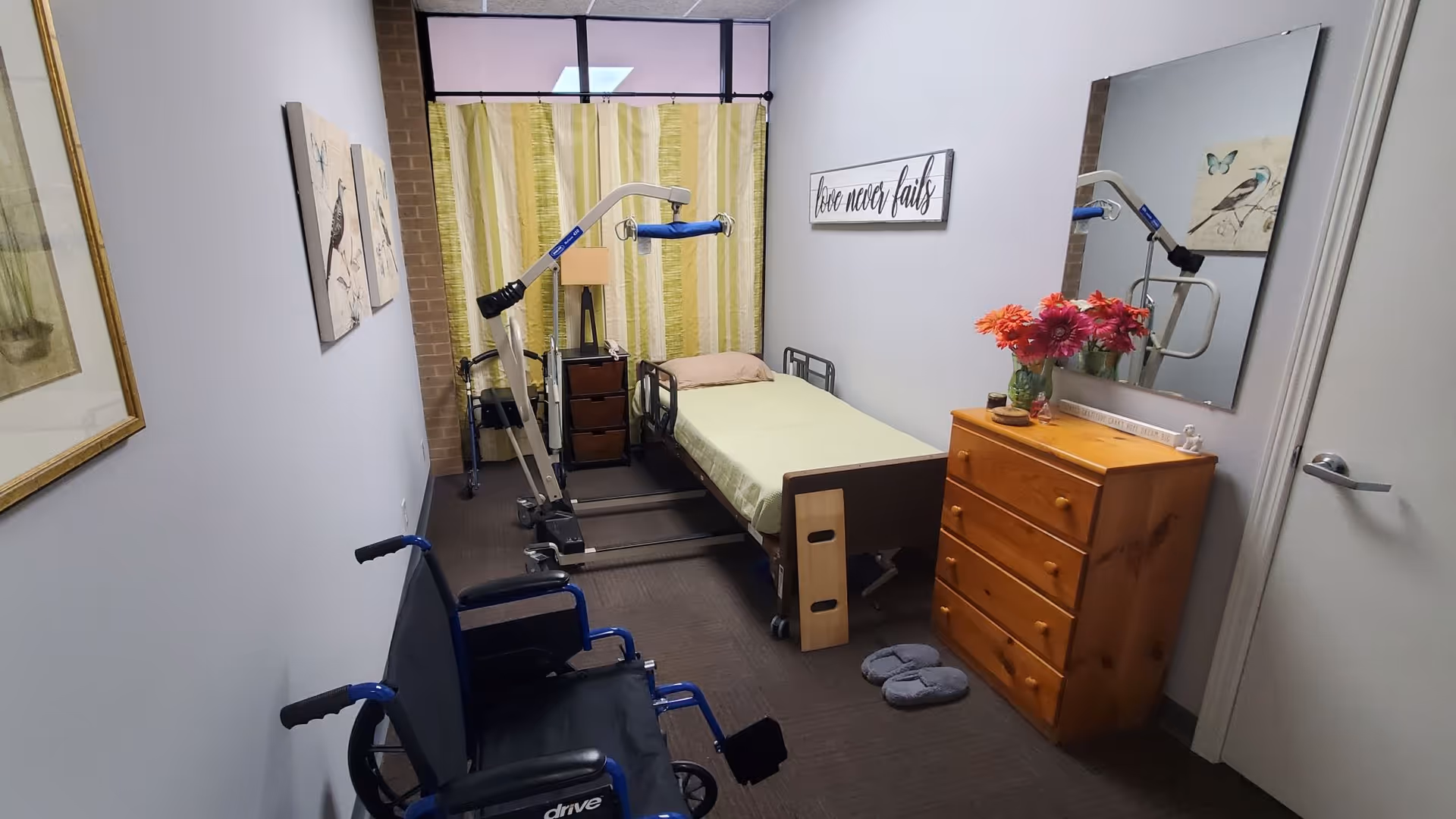 A small senior care bedroom with bed, lift device, wheelchair, dresser, mirror, and flowers.
